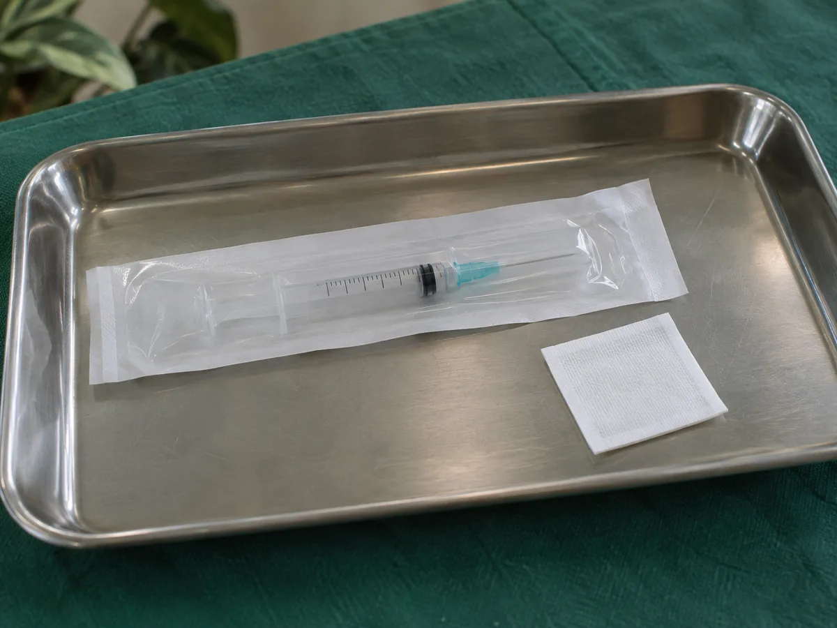 A sealed syringe package and a swab on a steel tray over a green drape
